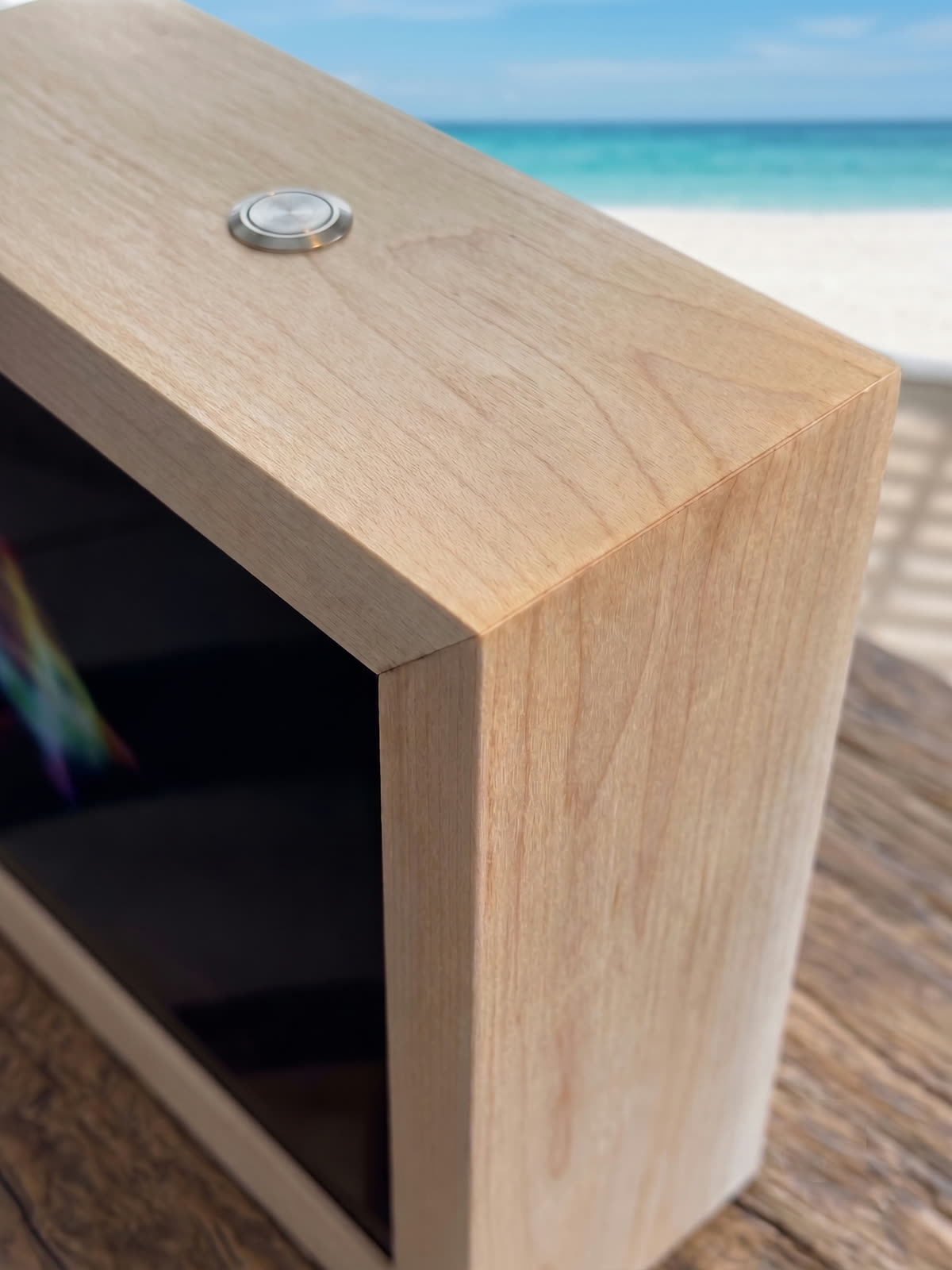 Close-up of the panel's top corner: a mitered maple frame with a clear finish, a stainless ask button set into the top, and the ocean out the window behind