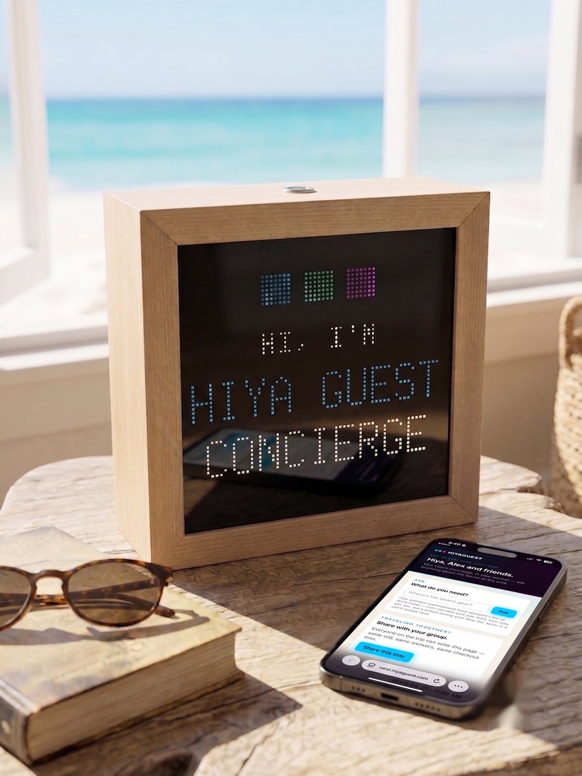 A wood-framed Hiya Concierge panel reading HI, I'M HIYA GUEST CONCIERGE beside a phone showing the stay's pocket concierge page, on a table in a beach rental